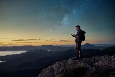 Person with backpack standing on a mountain at sunset, looking at phone. Stars in the sky.