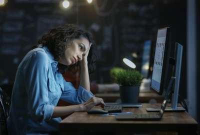 Woman in blue shirt, tired, working on a computer at night.